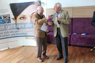 Zwei lächelnde ältere Männer; einer mit Mikrofon überreicht dem anderen ein Objekt vor einem Werbebanner.