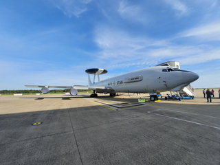 Ein großes, graues NATO E-3 Sentry AWACS Flugzeug mit charakteristischem Radom auf dem Rollfeld unter blauem Himmel.