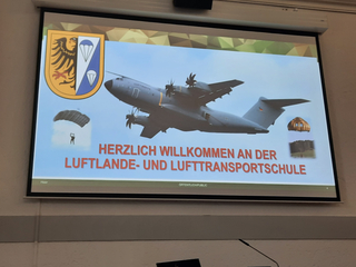 Willkommensfolie der Luftlande- und Lufttransportschule mit Militärflugzeug, Fallschirmspringer und Emblem.
