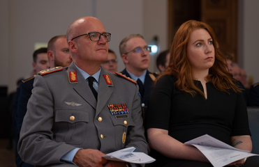 Ein Militäroffizier mit Medaillen und eine rothaarige Frau sitzen nebeneinander und beobachten aufmerksam ein Ereignis.