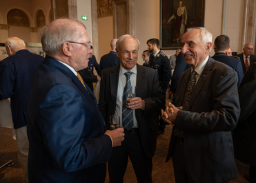 Drei ältere Männer in Anzügen lächeln und unterhalten sich. Einer hält ein Getränk, bei einer formellen Veranstaltung.