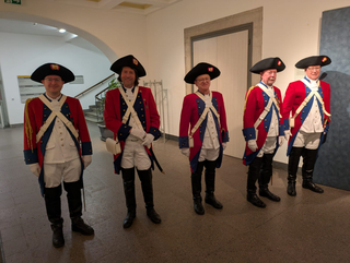 Fünf Männer in historischen rot-blauen Militäruniformen, weißen Kniehosen, schwarzen Stiefeln und Dreispitzen.