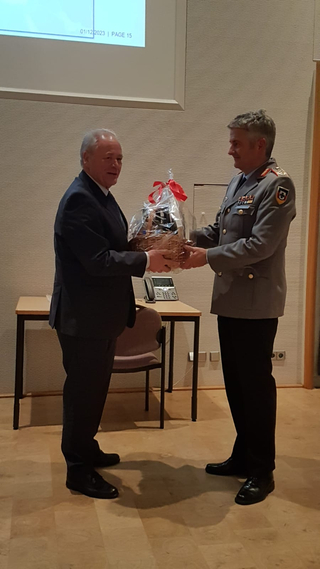 Ein Mann im dunklen Anzug überreicht einem lächelnden Mann in grauer Militäruniform einen verpackten Geschenkkorb.