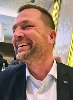 Nahaufnahme eines lächelnden Mannes mit Bart und kurzen Haaren, Anzug und Reversnadel, bei einer Veranstaltung.