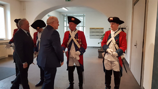 Männer in traditionellen roten Militäruniformen und Anzügen schütteln sich in einem modernen Flur die Hände.