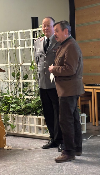 Zwei Männer, einer in grauer Militäruniform und einer in brauner Trachtenjacke, stehen nebeneinander bei einer Veranstaltung.