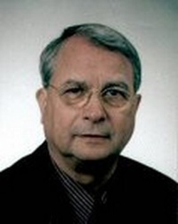 Kopfaufnahme eines älteren Mannes mit grauem Haar und Brille, der eine dunkle Anzugjacke über einem gestreiften Hemd trägt.