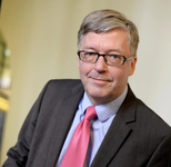 Professionelles Porträt eines Mannes mit grauem Haar, Brille, dunklem Anzug, hellblauem Hemd und rosa Krawatte, lächelnd.