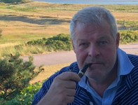 Mann mit grauem Haar und Bart, gestreiftem Hemd, eine Pfeife rauchend, mit Küstendünen und Meer im Hintergrund.