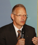 Ein Mann im Anzug mit Brille und Schnurrbart spricht in ein Mikrofon, offenbar bei einer Präsentation oder Rede.