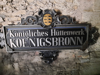 Verziertes Gusseisenschild an Steinmauer mit der Aufschrift „Königliches Hüttenwerk KOENIGSBRONN“ und Württembergischem Wappen.