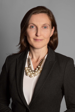 Professionelles Porträt einer Frau mit braunen Haaren und leichtem Lächeln, trägt einen dunklen Blazer und eine Perlenkette.