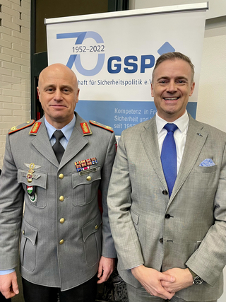 Ein Militäroffizier in Uniform und ein Mann im Anzug lächeln vor einem GSP-Banner zum 70-jährigen Jubiläum.