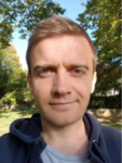 Lächelnder Mann mit kurzen braunen Haaren und blauen Augen, draußen vor Bäumen und blauem Himmel stehend.
