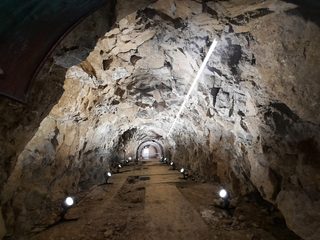 Ein hell beleuchteter, ausgegrabener Felstunnel mit rauen Wänden und einer langen diagonalen Lichtleiste, die zu einem gemauerten Bogen führt.
