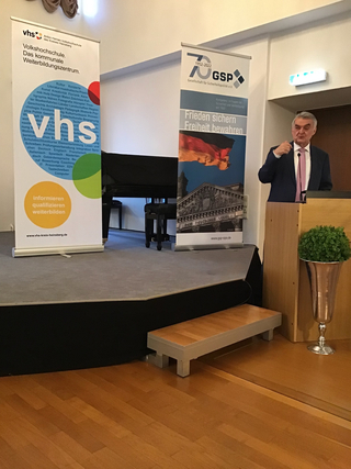 Mann spricht vor VHS Heinsberg und GSP-Bannern, die Bildung und Sicherheitspolitik thematisieren.