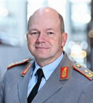 Porträt eines lächelnden, glatzköpfigen Mannes in grauer Militäruniform mit goldenen und roten Schulterstücken.