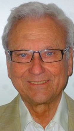 Porträt eines lächelnden älteren Mannes mit grauem Haar, blauen Augen und Brille.