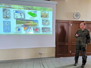 Ein Soldat in Uniform steht neben einem Bildschirm, der eine Präsentation über den Kampfpanzer Leopard 2 A7V und seine Upgrades zeigt.