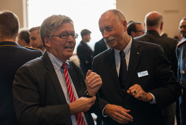 Zwei Männer in Anzügen unterhalten sich auf einer Veranstaltung. Der linke Mann mit Brille gestikuliert, der rechte lächelt.