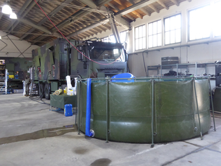 Ein getarnter Militär-LKW (Iveco) steht in einer Halle neben einem großen, runden olivgrünen flexiblen Wassertank.