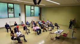 Eine Gruppe von Personen, einige mit Masken, sitzen verteilt in einem Raum und hören einem uniformierten Mann zu.