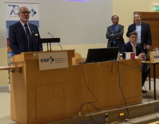 Ein bärtiger Mann spricht am Rednerpult mit GSP-Logo vor Publikum, weitere Männer stehen und sitzen im Hintergrund.