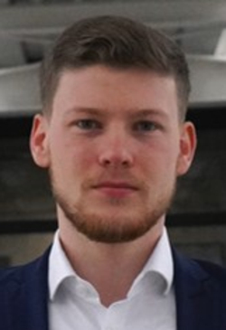 Ein junger Mann mit Bart, einem dunkelblauen Anzug und weißem Hemd steht in einem modernen Gebäude und blickt in die Kamera.