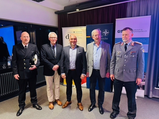 Fünf Männer, darunter zwei in Bundeswehr-Uniformen (Marine und Heer), stehen vor Konferenzbannern für ein Gruppenfoto.