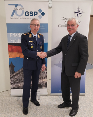 Zwei Männer, einer in Uniform und einer im Anzug, schütteln sich die Hände vor Bannern der GSP und Deutschen Atlantischen Gesellschaft.