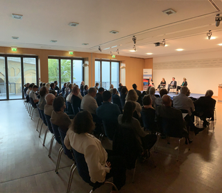 Zuschauer in einem Konferenzraum verfolgen eine Podiumsdiskussion mit Referenten auf der Bühne. Große Fenster links.