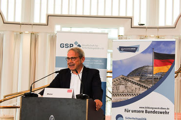 Mann (Maier) spricht am Rednerpult vor Bannern der Gesellschaft für Sicherheitspolitik und des Bildungswerks mit Reichstag und deutscher Flagge.