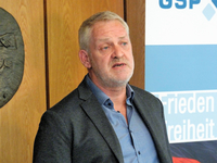 Mann mit grauem Haar und Bart spricht vor einem Banner mit den teilweise sichtbaren Worten "Frieden" und "Freiheit".