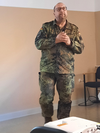 Soldat der Bundeswehr in Flecktarn-Uniform, Brille und Bart, steht mit gefalteten Händen in einem Raum.