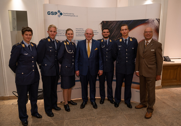 Sieben Personen, sechs in Uniform, und ein älterer Mann im Anzug, posieren vor einem GSP-Banner.