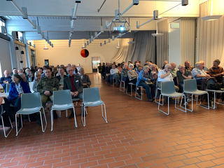 Ein Raum mit vielen sitzenden Menschen bei einer Veranstaltung oder Präsentation, einige Stühle im Vordergrund sind leer und reserviert.