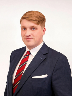 Porträt eines lächelnden jungen Mannes mit rötlich-blondem Haar, dunklblauem Anzug, weißem Hemd und gestreifter Krawatte.