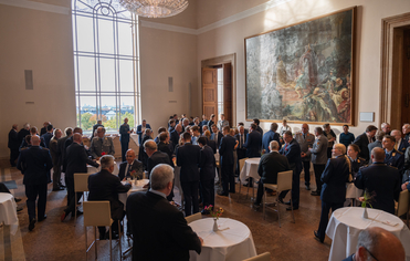 Eine große Gruppe von Menschen, viele in Uniform, unterhält sich in einem großen Saal mit hohen Decken, großen Fenstern und einem Gemälde.