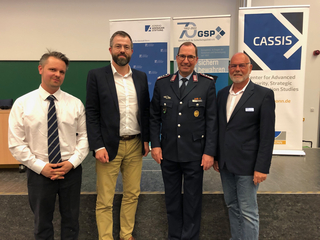 Vier lächelnde Männer, darunter ein Uniformierter, vor Bannern der Konrad-Adenauer-Stiftung, GSP und CASSIS.