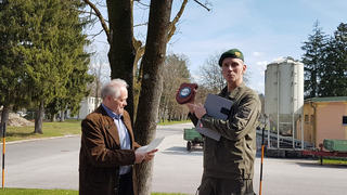 Ein Offizier in Uniform überreicht einem älteren Zivilisten, der Papiere hält, im Freien eine schildförmige Auszeichnung.
