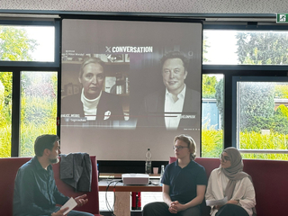 Drei Personen verfolgen eine Projektion, die eine 'X CONVERSATION' zwischen Alice Weidel und Elon Musk zeigt.