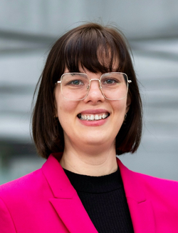 Lächelnde Frau mit kurzen braunen Haaren, goldener Brille und leuchtend pinkfarbenem Blazer, auf einer grauen Leiste lehnend.