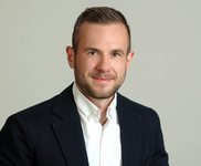 Ein lächelnder junger Mann mit kurzen braunen Haaren und Bart, trägt einen dunklen Blazer und weißes Hemd, blickt in die Kamera.
