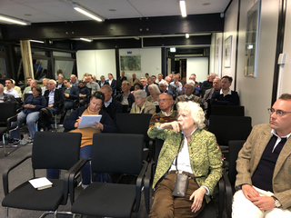 Publikum aus verschiedenen Erwachsenen und Senioren sitzt in Reihen in einem Konferenzraum und nimmt an einem Vortrag oder Treffen teil.