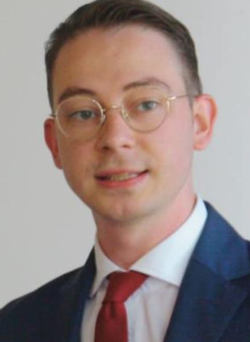 Ein junger Mann im blauen Anzug, roter Krawatte und Brille lächelt an einem Rednerpult mit Mikrofonen.
