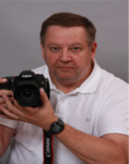 Ein Mann in weißem Poloshirt hält eine Canon Spiegelreflexkamera mit einem ernsten Blick vor grauem Hintergrund.