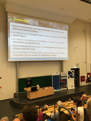 Militäroffizier präsentiert im Hörsaal vor Publikum über Weltraum-Sicherheit und -Strategie. Bildschirm zeigt 'Kernbotschaften'.