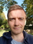 Porträt eines lächelnden Mannes mit kurzen braunen Haaren und blauen Augen, in blauem Hoodie, draußen mit Bäumen und blauem Himmel.