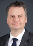 Professionelles Porträt eines Mannes mit grauem Haar und braunen Augen, dezent lächelnd, im dunklen Anzug mit Krawatte und Anstecknadel.