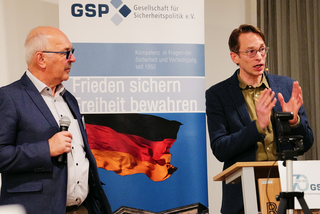 Zwei Männer bei einer GSP-Veranstaltung. Einer spricht am Rednerpult, der andere hält ein Mikrofon. Deutscher Flaggenhintergrund.